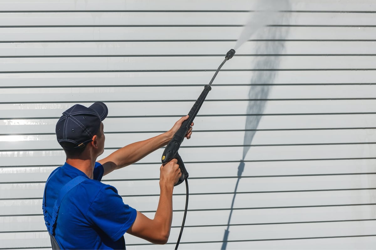 Worker in overalls washes a white wall from siding with a water gun, cleaning the facade of a house