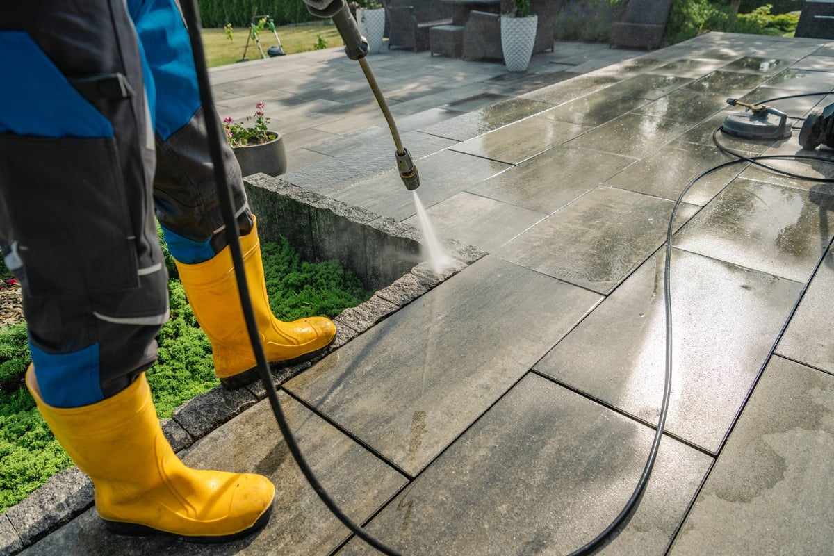A worker in yellow boots cleans a stone patio with a pressure washer on a sunny day