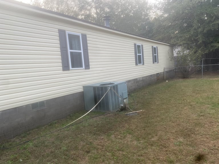 Single-wide mobile home with white siding and blue-trimmed windows, surrounded by grass and trees
