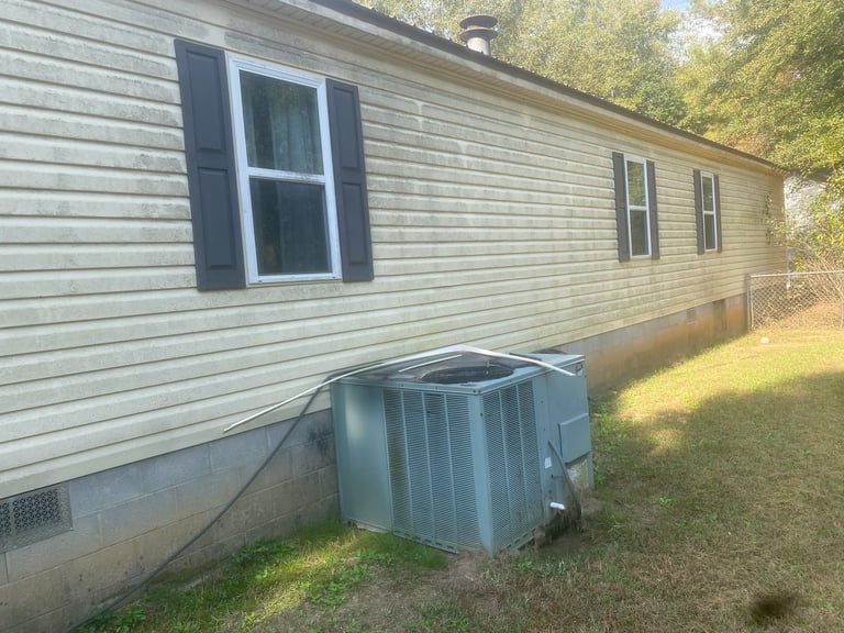 Single-wide mobile home exterior with beige siding, blue-trimmed window, and metal HVAC unit on grass ground