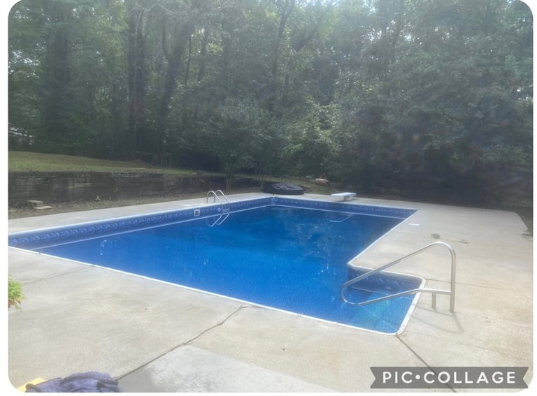 Empty rectangular blue swimming pool surrounded by concrete deck and lounge chair, with dense green trees in background