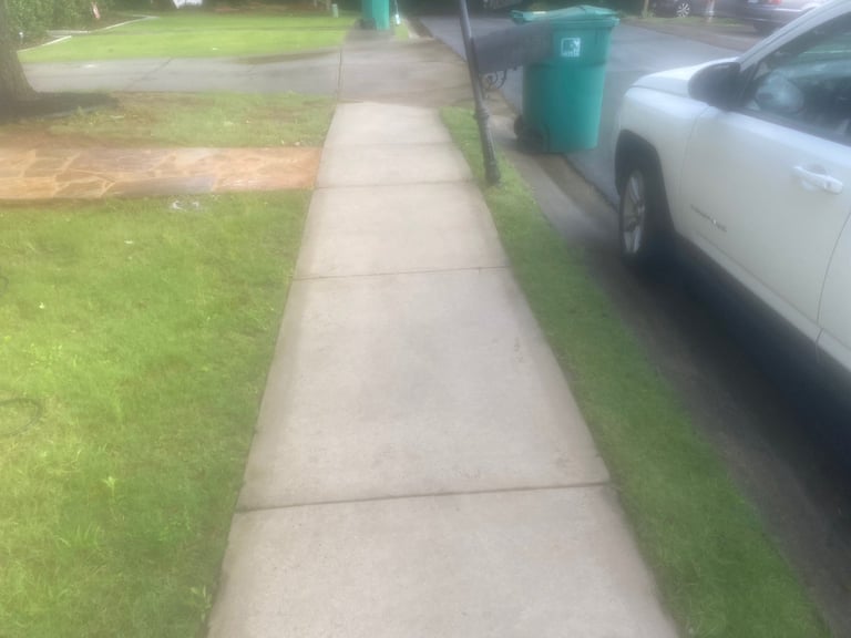 Concrete sidewalk running alongside a residential street with green grass, white car, and trash bin visible