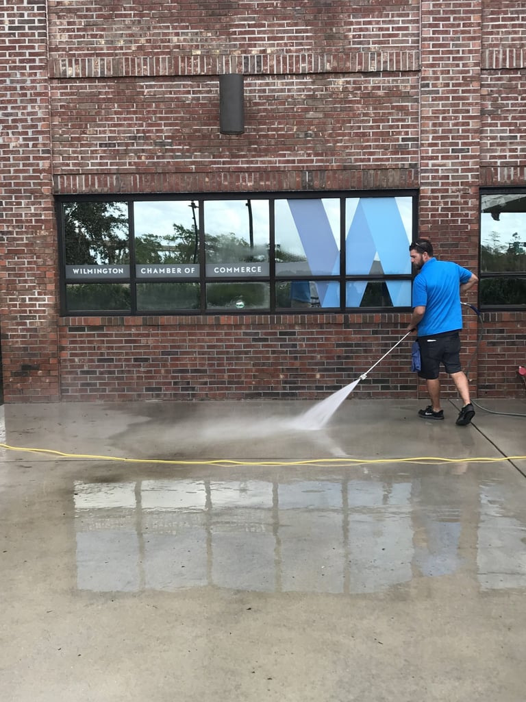 Man in blue shirt power washing concrete pavement in front of brick building storefront windows